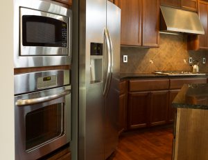 Closeup photo of stainless steel appliances in modern residential kitchen with stone counter tops and cherry wood cabinets with hardwood floors